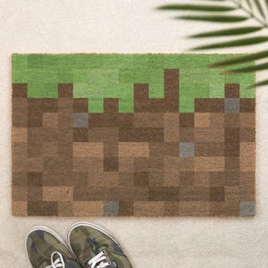 May include: A rectangular doormat featuring a pixelated block pattern with a green top section representing grass and a brown lower section representing dirt, accented with small grey squares throughout the design, laid on a light surface.