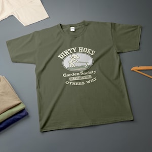 May include: An olive green short-sleeve t-shirt featuring a graphic of a person using a hoe in a garden. The text reads: DIRTY HOES, Garden Society, WE THRIVE WHERE OTHERS WILT. The design is printed in a light cream colour on the centre of the chest.