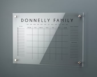 May include: A clear acrylic dry-erase board with a white background and black text. The board has a calendar grid with the months of the year, days of the week, and a to-do list. The text "DONNELLY FAMILY" is at the top of the board.