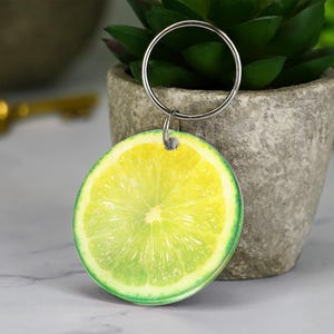 May include: A round keychain featuring a realistic image of a lime slice. The keychain has a silver metal ring attached to the top. The lime slice is yellow and green, with a detailed texture.