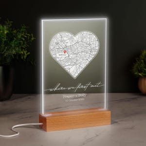 May include: An illuminated map display with a heart-shaped design, set on a wooden base. The map shows a location marked with a red pin. Text reads "where we first met", names, and a date. The light emits a white glow.