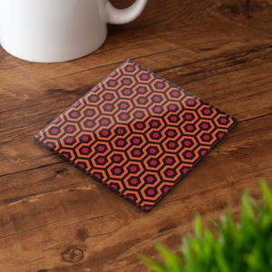 May include: Square coaster featuring a repeating geometric pattern in orange, black, and burgundy. The coaster is designed to protect surfaces from drink condensation. A white mug is in the background. The coaster measures approximately 10 cm x 10 cm.