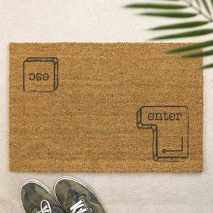 May include: A rectangular brown coir doormat featuring the words "esc" and "enter" in black, styled like computer keyboard keys. The mat rests on a light surface, with camouflage sneakers at the bottom.