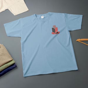 K&ouml;nnte beinhalten: Hellblaues T-Shirt mit einem Grafikdesign auf der Vorderseite. Das Design ist in Rot, Schwarz und Grau gehalten, mit dem Wort "TRIUMPH" darunter. Mehrere gefaltete Hemden sind unten links im Bild zu sehen.