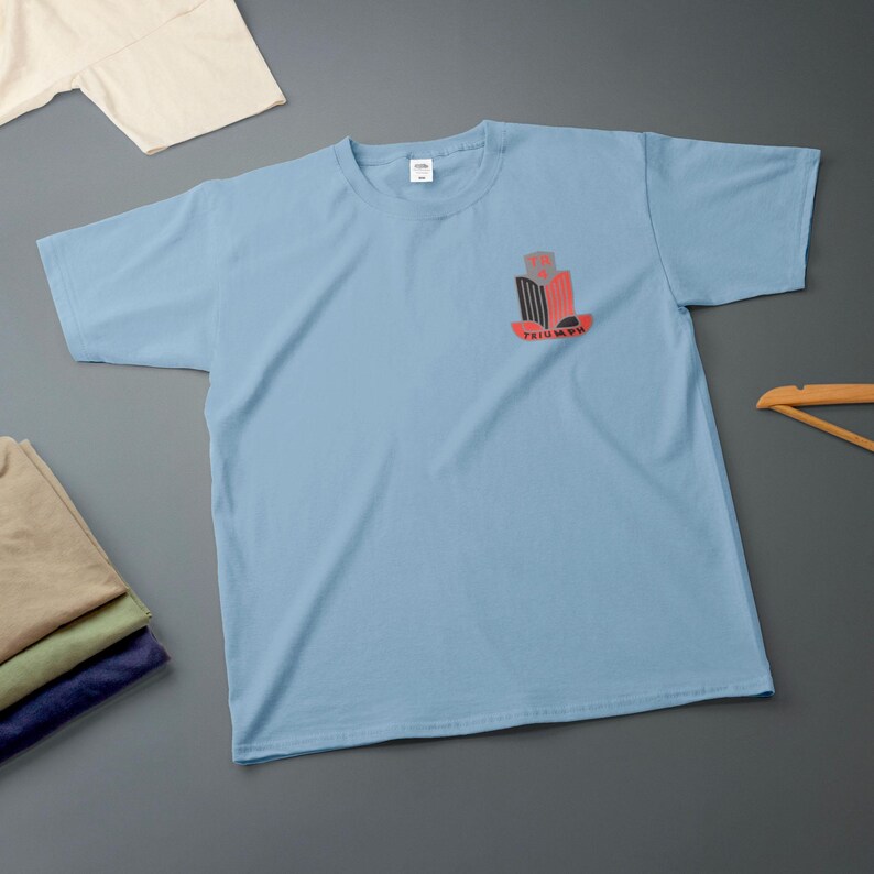 K&ouml;nnte beinhalten: Hellblaues T-Shirt mit einem Grafikdesign auf der Vorderseite. Das Design ist in Rot, Schwarz und Grau gehalten, mit dem Wort "TRIUMPH" darunter. Mehrere gefaltete Hemden sind unten links im Bild zu sehen.