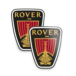 May include: Two black and gold Rover logo stickers. Each sticker features a shield shape with the word "ROVER" at the top. Below is a Viking ship design with red sails and gold accents. The stickers have a white border.