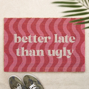 May include: A rectangular doormat with a wavy pattern in shades of red and pink. The text "better late than ugly" is printed in a light beige color across the center. A pair of camouflage sneakers are at the bottom.