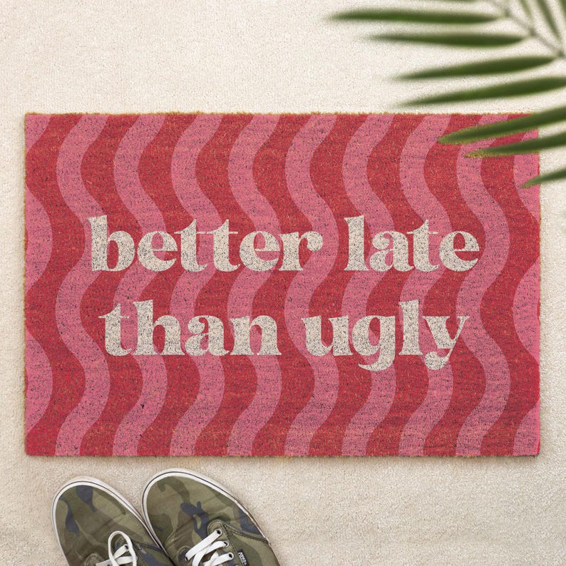 May include: A rectangular doormat with a wavy pattern in shades of red and pink. The text "better late than ugly" is printed in a light beige color across the center. A pair of camouflage sneakers are at the bottom.