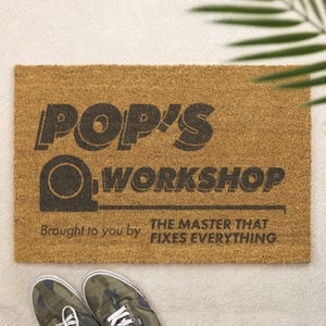 May include: A rectangular coir doormat in natural brown. It displays the text "POP'S WORKSHOP" in dark grey, with a tape measure graphic. The text below reads "Brought to you by THE MASTER THAT FIXES EVERYTHING".