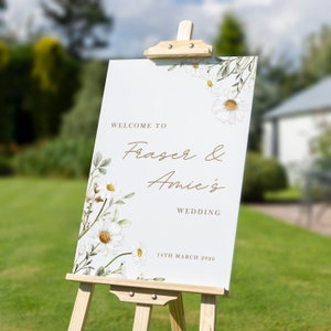 Op de afbeelding: Een wit welkomsbord voor een bruiloft met een bloemenontwerp met witte madeliefjes en groene bladeren. Het bord staat er: "Welcome to Fraser & Amie's Wedding 14th March 2026".