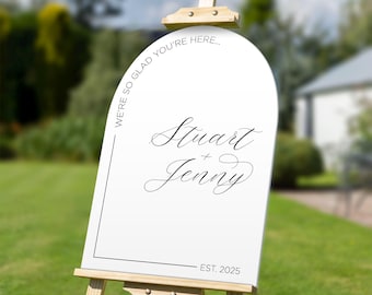 Puede incluir: Un letrero de bienvenida de boda blanco sobre un caballete de madera. El letrero tiene el texto "We're so glad you're here..." y los nombres "Stuart + Jenny" en escritura elegante, con "EST. 2025" en la parte inferior. El fondo es un césped verde.
