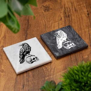 May include: Two square coasters with a raven perched on a skull design. One coaster is white with a black raven and skull, the other is dark gray with a white raven and skull. The coasters are on a wooden surface.