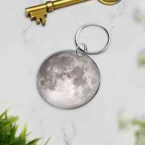 Può includere: Un portachiavi rotondo con un'immagine dettagliata della luna. La superficie lunare è raffigurata in tonalità di grigio e bianco, con crateri e texture visibili. Il portachiavi ha un anello color argento per attaccare le chiavi.