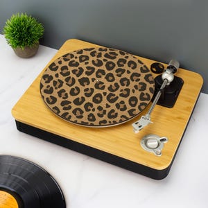 Puede incluir: Un tocadiscos de madera con un disco con estampado de leopardo. El tocadiscos tiene un acabado de madera marrón claro con una base negra. El brazo y los controles son plateados. Una pequeña planta en maceta está en el fondo.