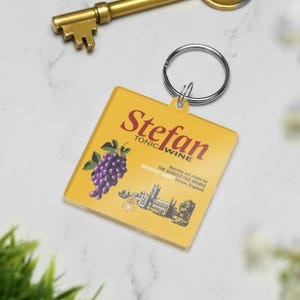May include: A yellow square keychain with the name "Stefan" in burgundy script, with "TONIC WINE" below. A purple grape illustration and text about Buckfast Abbey are also featured. A gold key is in the background.