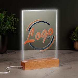Op de afbeelding: Een rechthoekige LED-lamp met een houten voet. Het licht heeft een logo-ontwerp in oranje en marineblauw met de tekst "Logo HERE". Het licht is ingeschakeld en de randen gloeien wit. Een witte kabel loopt vanaf de voet.