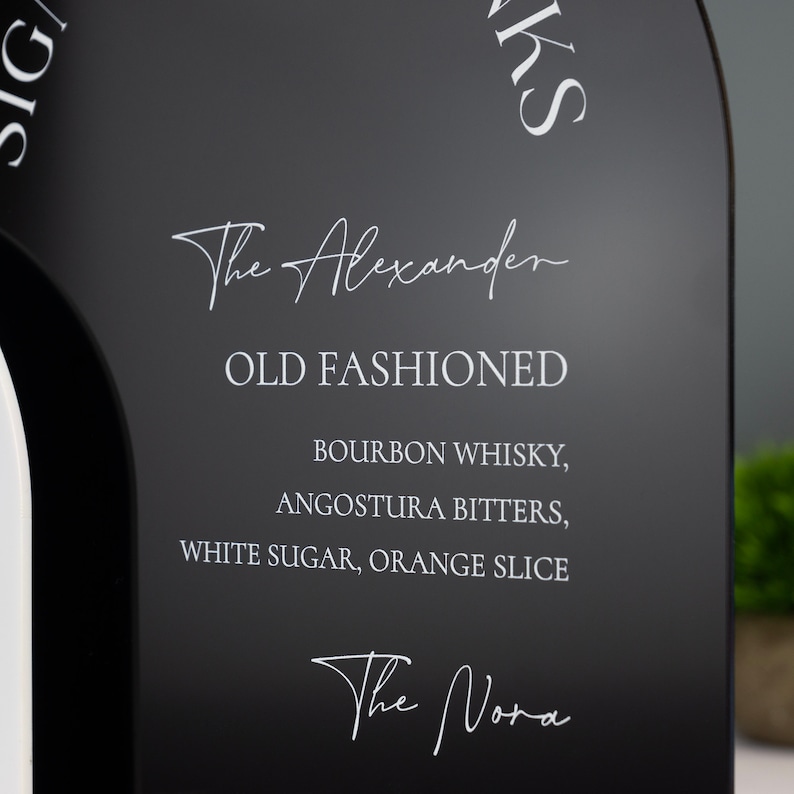 K&ouml;nnte beinhalten: Schwarzes Schild mit wei&szlig;em Text, der die Zutaten f&uuml;r einen Old Fashioned Cocktail auflistet. Der Text lautet "The Alexander", "Old Fashioned", "Bourbon Whiskey, Angostura Bitters, White Sugar, Orange Slice" und "The Nora".