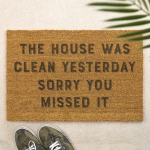 Op de afbeelding: Een rechthoekige deurmat van natuurlijke kokosvezel, met de tekst "THE HOUSE WAS CLEAN YESTERDAY SORRY YOU MISSED IT" in zwarte letters. Twee sneakers met camouflageprint staan onderaan.