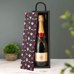 Puede incluir: Una botella de champán en una caja de regalo de madera negra. El interior de la caja es negro y la tapa presenta un patrón de corazones rosas, calaveras y llamas sobre un fondo gris oscuro. La botella tiene una parte superior de lámina dorada y un sello rojo.