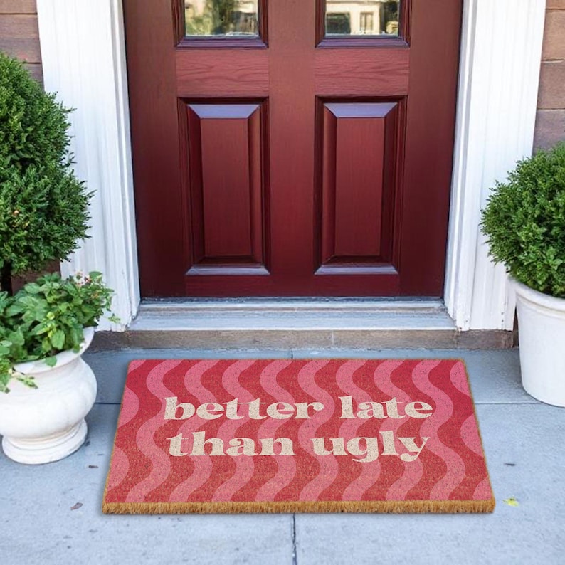 May include: A rectangular doormat with a wavy pattern in shades of red and pink. The text "better late than ugly" is displayed in a light beige color. The mat is placed in front of a dark red door.
