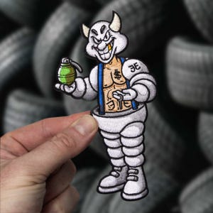 Puede incluir: Un parche bordado que muestra un personaje de dibujos animados blanco con cuernos, torso musculoso con un signo de dólar y tirantes azules. El personaje sostiene una granada verde y tiene las letras 3E en el hombro. El diseño está bordeado en negro.