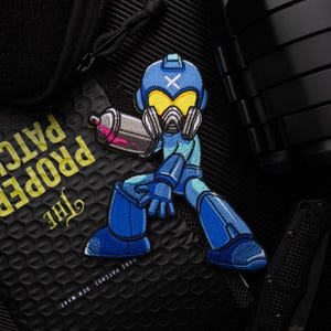 May include: A blue and yellow embroidered patch of a robot character holding a spray can. The robot is wearing a helmet with a cross on it and a gas mask. The patch is on a black background.
