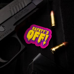 Peut inclure: Un pistolet noir avec le texte "SIG SAUER INC" et deux balles. Un écusson coloré avec les mots "SAFETY'S OFF!" en jaune et un bord rose est également visible.