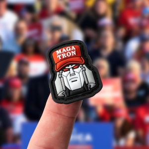 May include: A rubberized patch featuring a stylized robot head in shades of gray and black, wearing a bright red baseball cap with the white text MAGA TRON printed on the front. The robot has sharp, angular features and narrow red eyes.