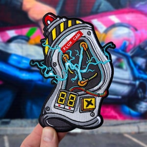 以下が含まれることがあります： フラックスキャパシタのデザインが施された刺繍パッチ。パッチは主にシルバー、ブラック、イエローで、青い稲妻と「FLUX CAN」の文字が特徴です。また、「88」と表示された2つのデジタルディスプレイも備えています。