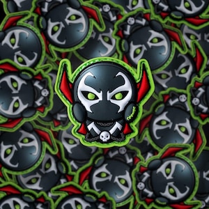May include: A collection of cartoon-style Spawn character patches. The patches feature a black and white face with green eyes, a red cape, and a skull necklace, outlined in green. The background is filled with similar, blurred patches.