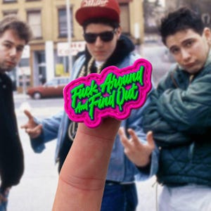 May include: A bright pink rubberized patch with a scalloped edge featuring the neon green text "Fuck Around And Find Out" in a stylized script font. The patch is held up by a finger, displaying a bold, high-contrast design with raised lettering.
