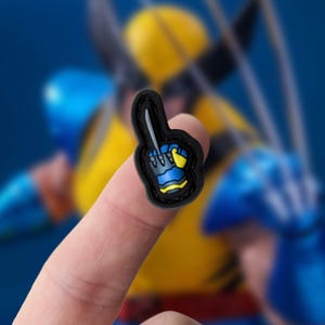 Puede incluir: Un títere de dedo negro y azul con una garra amarilla y una cuchilla plateada. El títere tiene la forma de una mano con el dedo índice extendido.