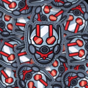May include: A close-up of a gray and red Ant-Man character patch. The patch is made of a soft, textured material and has a stitched border. The character's face is visible, with red eyes and a white stripe across the forehead.