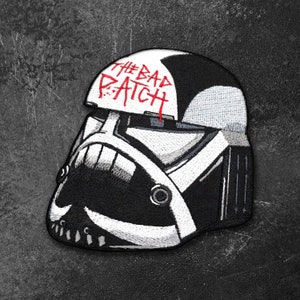 May include: An embroidered patch featuring a stylized black and white helmet design. The top section of the helmet displays the text THE BAD PATCH in red lettering. The design includes bold black and white geometric shapes and lines.