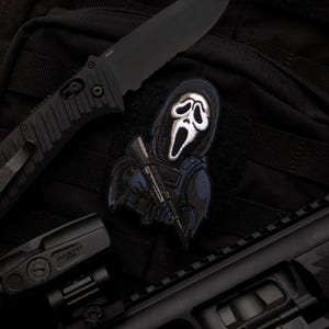 Può includere: Toppa tattica nera con il personaggio di Ghostface che tiene un fucile, con un coltello nero e un'arma da fuoco sullo sfondo. La toppa ha una faccia bianca e un cappuccio nero, con dettagli blu e neri. Su sfondo nero.