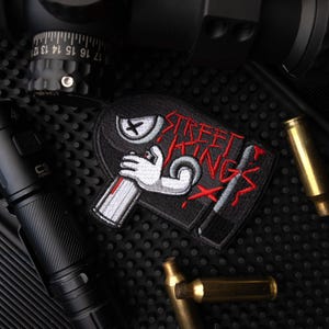 May include: A black embroidered patch featuring a cartoon hand holding a silver spray paint can. The design includes a stylized eye with an X for a pupil and the red text STREET KINGS. The patch has a black border and is shaped like a spray can.