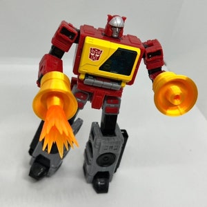 May include: A red and gray robot toy with a yellow chest plate and a black Autobot symbol. The robot is holding two orange and yellow blasters in each hand. One blaster is firing a bright orange spark.