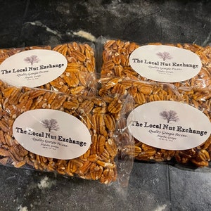 May include: Four clear plastic bags filled with pecan halves. Each bag has a white oval label with the text "The Local Nut Exchange - Quality Georgia Pecans" and a tree graphic. The pecans are a light brown color.