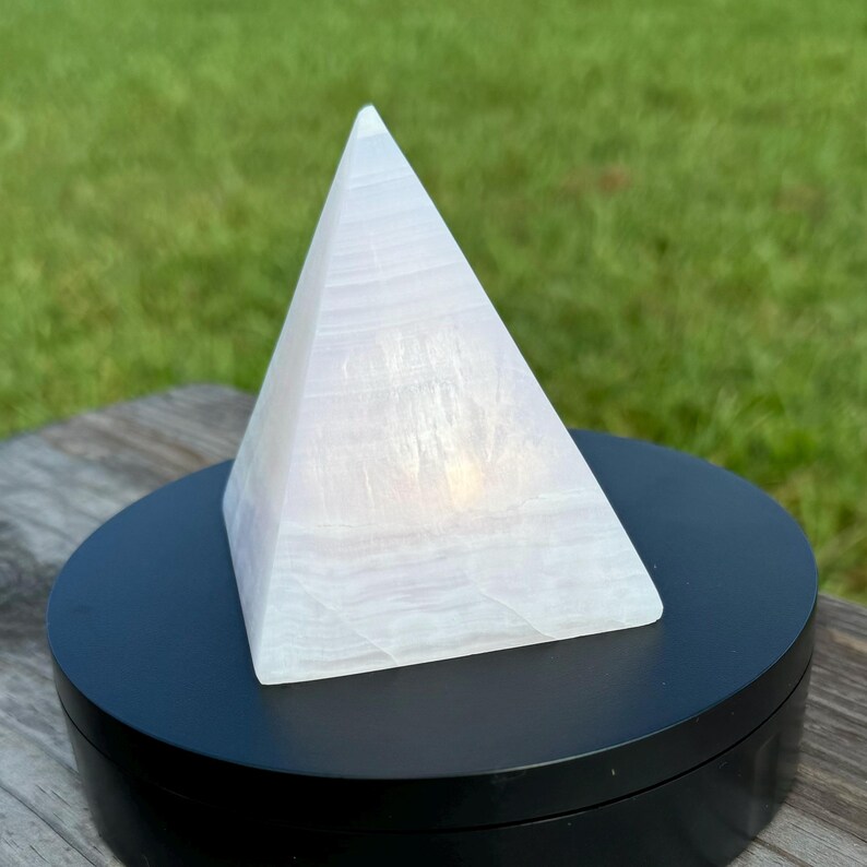 Op de afbeelding: Een witte selenietkristalpiramide op een zwarte ronde basis. De piramide is glad en doorschijnend, met een lichte glans.