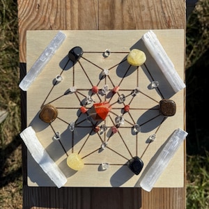 May include: A wooden Metatron's Cube grid with various gemstones. The grid features a geometric design with a central red crystal, surrounded by clear, yellow, brown, and black stones. White selenite wands are placed around the grid's perimeter.