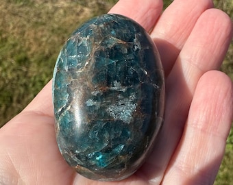 Op de afbeelding: Een gepolijste, ovaalvormige apatiet-edelsteen met diepe blauwgroene tinten, donkerbruine minerale insluitingen en interne breuken over het gehele oppervlak, met een gladde, glanzende textuur en een reflecterende kristalstructuur.