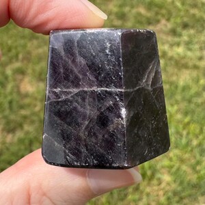 May include: A polished, dark purple, trapezoidal-shaped stone with visible internal fractures and a smooth, reflective surface. The mineral displays deep violet hues with lighter crystalline inclusions throughout its structure, showcasing a geometric cut.