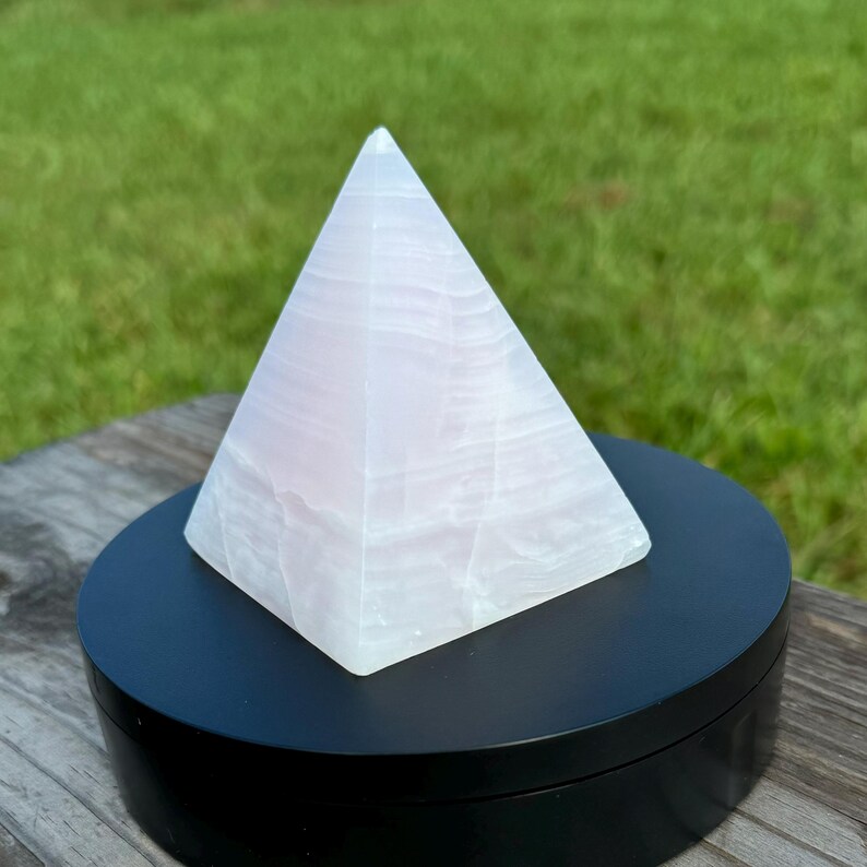 Op de afbeelding: Een witte seleniet piramidekristal op een zwarte displaystandaard. De piramide is glad en heeft een doorschijnende uitstraling.