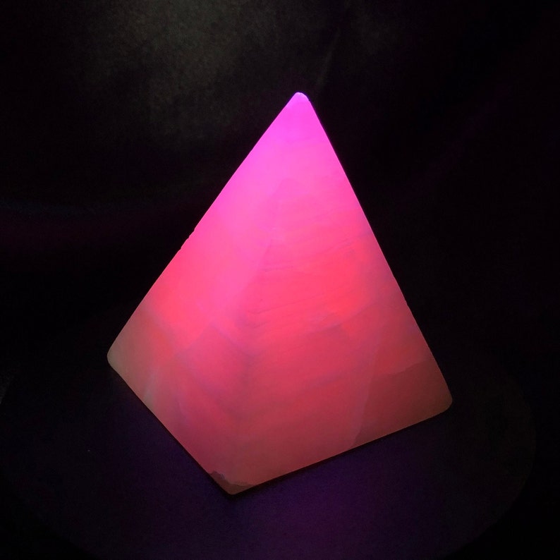 Op de afbeelding: Een roze kristallen piramide met een glad, gepolijst oppervlak. De piramide wordt van binnenuit verlicht, waardoor een zachte roze gloed ontstaat.
