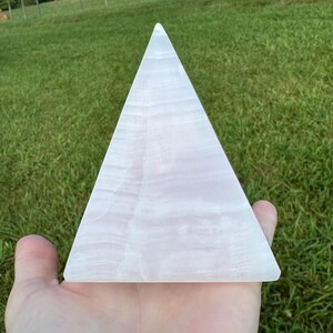 Op de afbeelding: Een witte seleniet piramidekristal. De piramide is glad en gepolijst, met een doorschijnende uitstraling. De kristal ligt op een hand.
