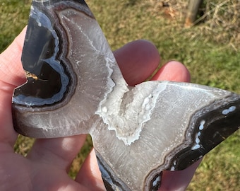 Op de afbeelding: Een vlindervormige plak agaatsteen met concentrische banden van donkerbruin, doorschijnend grijs en witte kristallijne patronen. Het gepolijste oppervlak toont natuurlijke minerale lagen met een grillig, sprankelend wit kwartscentrum.