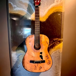 Può includere: Una chitarra acustica in legno con finitura marrone è esposta in una cornice di vetro. La chitarra presenta diverse firme e scritte sul corpo. La cornice ha uno sfondo blu chiaro e giallo.
