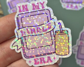 Può includere: Adesivo olografico con un e-reader rosa con la scritta "IN MY SINGLE ERA" su uno striscione. Una piccola bevanda con una cannuccia è accanto all'e-reader. L'adesivo ha una finitura glitterata e iridescente.