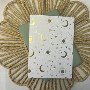Könnte beinhalten: Ein weißes Notizbuch mit einem Himmelsdesign mit goldenen Monden, Sternen und Sonnen. Das Notizbuch liegt auf einem salbeigrünen Einband, auf einem gewebten, kreisförmigen Platzset.