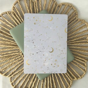 Peut inclure: Une carte rectangulaire avec un motif céleste, ornée de constellations dorées, de lunes et d'étoiles sur un fond gris clair. La carte repose sur un objet vert sauge, posé sur un dessous de plat circulaire tressé.